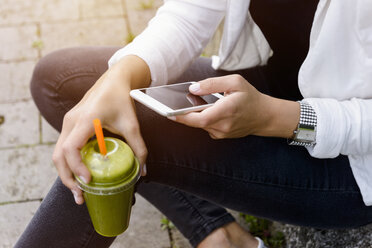 Abgeschnittene Aufnahme einer jungen Frau, die mit einem Smoothie-Getränk sitzt und ein Smartphone benutzt - CUF30176