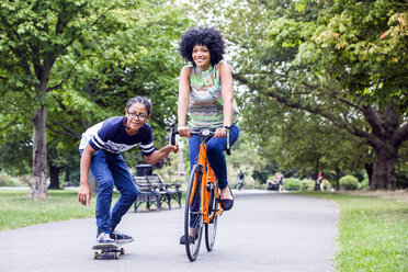 Skateboard fahrender Junge hält sich im Park am Fahrrad seiner Mutter fest - CUF30239