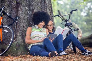 Mature woman and son sitting against park tree reading book and digital tablet - CUF30242