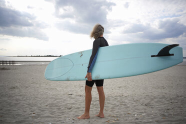 Ältere Frau steht am Strand und hält ein Surfbrett, Rückansicht - CUF30251