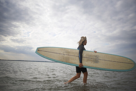 Ältere Frau geht vom Meer weg, trägt Surfbrett, lizenzfreies Stockfoto