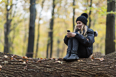 Mädchen benutzt Smartphone auf Baumstamm im Herbstwald - CUF30524