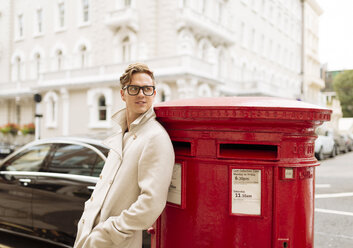 Stylish young man leaning against red post box, London, England, UK - CUF30777