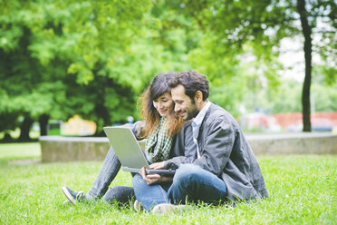 Junges Paar, das im Schneidersitz im Gras sitzt und einen Laptop benutzt und lächelnd nach unten schaut - CUF30854