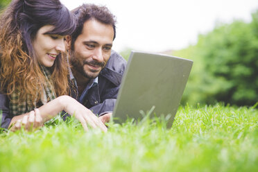 Kopf und Schultern eines jungen Paares, das auf der Vorderseite im Gras liegt und lächelnd einen Laptop benutzt - CUF30856