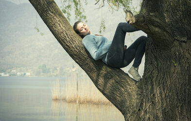 Young woman reclining in tree on lakeside, Lake Mergozzo, Verbania, Piemonte, Italy - CUF31124