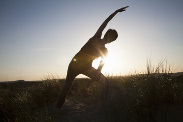 Rear view of mid adult woman practicing yoga in silhouetted long grass at sunset - CUF31191