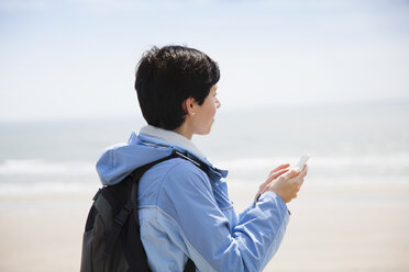 Mid adult woman texting on smartphone from beach while looking out to sea - CUF31193