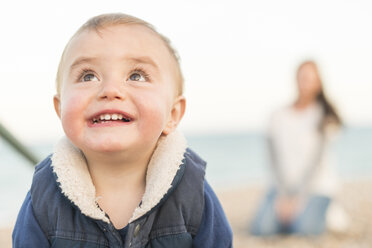 Porträt eines kleinen Jungen am Strand mit einer dahinter sitzenden Mutter - CUF31248