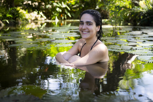 Junge Frau umgeben von Seerosenblättern im Teich sitzend, die Knie umarmend, lächelnd in die Kamera schauend - CUF31256