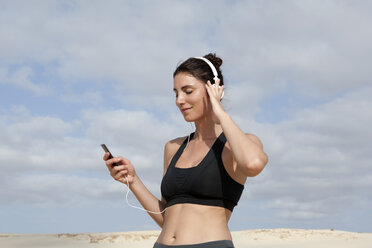 Mittlere erwachsene Frau wählt Musik für Kopfhörer aus, während sie am Strand trainiert - CUF31320