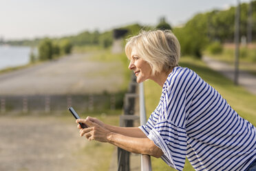 Smiling senior woman with cell phone leaning on railing at riverbank - FMKF05146