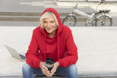 Ältere Frau mit rotem Kapuzenpulli sitzt im Freien mit Laptop und Kaffee zum Mitnehmen - FMKF05152