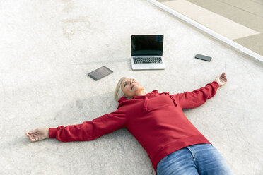 Senior woman wearing red hoodie lying on the ground next to mobile devices - FMKF05155