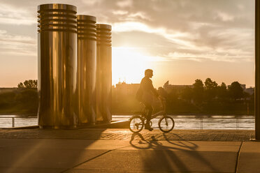 Senior woman riding city bike at the riverside at sunset - FMKF05175