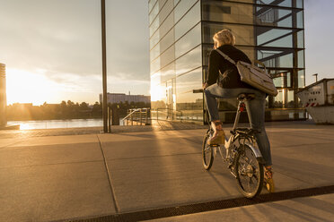 Senior woman with city bike at the riverside at sunset - FMKF05177