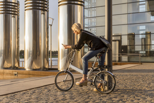 Senior woman with city bike using cell phone at the riverside at sunset - FMKF05182