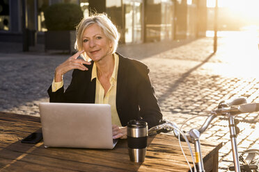 Lächelnde ältere Geschäftsfrau mit Laptop in der Stadt bei Sonnenuntergang - FMKF05184