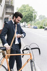 Lächelnder Geschäftsmann mit Fahrrad auf der Straße, der die Zeit kontrolliert - UUF14090