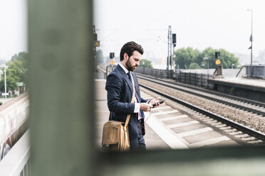 Geschäftsmann mit Mobiltelefon und Kopfhörern wartet am Bahnsteig - UUF14103