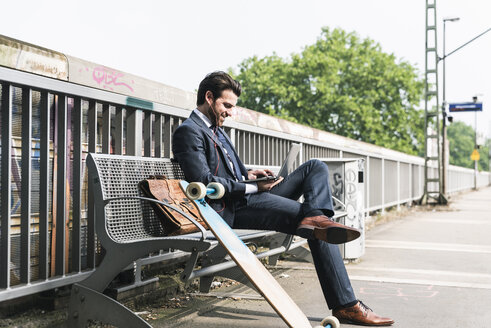 Lächelnder Geschäftsmann mit Skateboard und Laptop auf dem Bahnsteig - UUF14114