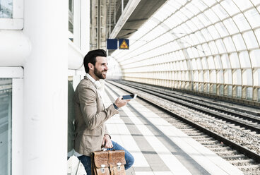Lächelnder junger Mann mit Mobiltelefon auf dem Bahnsteig - UUF14131