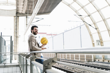 Lächelnder junger Mann mit Handy und Basketball auf dem Bahnsteig - UUF14135