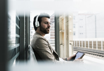 Young man with closed eyes wearing headphones and holding tablet at the station platform - UUF14143