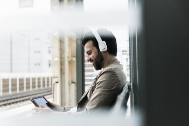 Smiling young man wearing headphones using tablet at the station platform - UUF14144