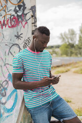 Young man in skatepark leaning against graffiti wall, listening music - ACPF00010