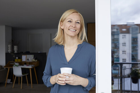 Smiling woman holding cup of coffee looking out of balcony door - RBF06299