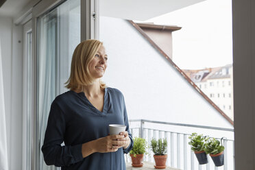 Lächelnde Frau mit einer Tasse Kaffee in der Hand und Blick aus der Balkontür - RBF06300