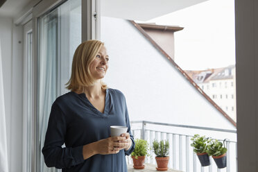 Smiling woman holding cup of coffee looking out of balcony door - RBF06300