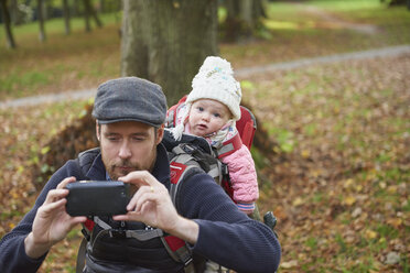 Mid adult man in park wearing flat cap carrying daughter on back in baby carrier taking selfie using smartphone - CUF31605