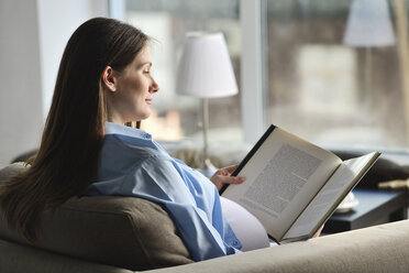 Schwangere Frau sitzt auf dem Sofa und liest ein Buch - CUF31740