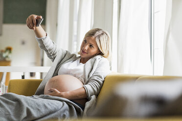 Hochschwangere junge Frau auf dem Sofa, die ein Selfie macht - CUF31789