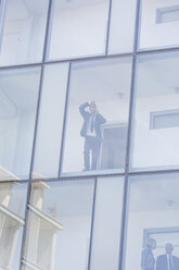 Geschäftsmann schaut durch das Fenster eines Hotels mit Glasfront, während er mit seinem Smartphone spricht - ISF09784