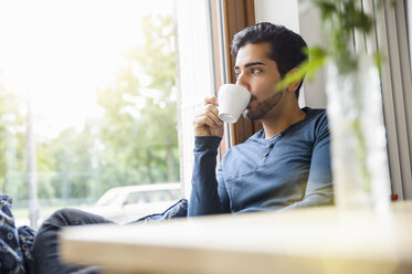 Junger Mann sitzt vor einem Fenster, trinkt Kaffee und schaut weg - CUF31861