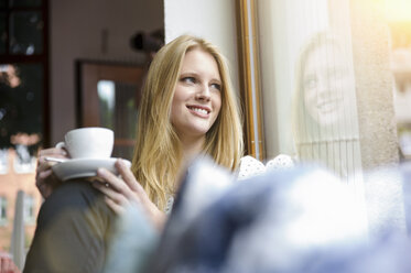 Junge Frau mit langen blonden Haaren sitzt mit Kaffeetasse in der Hand und schaut lächelnd weg - CUF31862