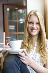 Junge Frau mit langen blonden Haaren sitzt mit einer Kaffeetasse in der Hand und schaut lächelnd in die Kamera - CUF31863