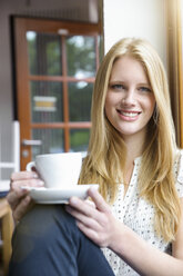 Young woman with long blond hair sitting holding coffee cup, looking at camera smiling - CUF31863