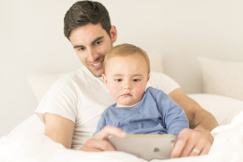 Vater und kleiner Sohn sitzen zusammen und schauen auf ihr Smartphone - CUF31897