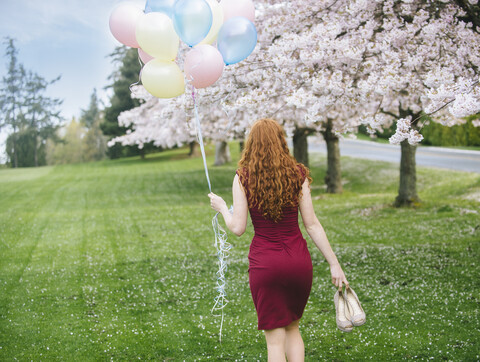 Rear view of young woman with long wavy red hair and bunch of balloons strolling in spring park stock photo