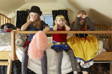 Three sisters, sitting on bed, wearing fancy dress costumes, covering mouth, ears and eyes - ISF10136