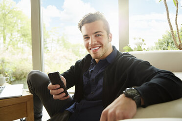 Portrait of smiling man sitting on floor holding smartphone - ISF10187