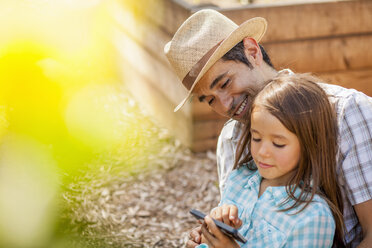 Mädchen auf dem Schoß des Vaters mit Smartphone im Gemeinschaftsgarten - ISF10305