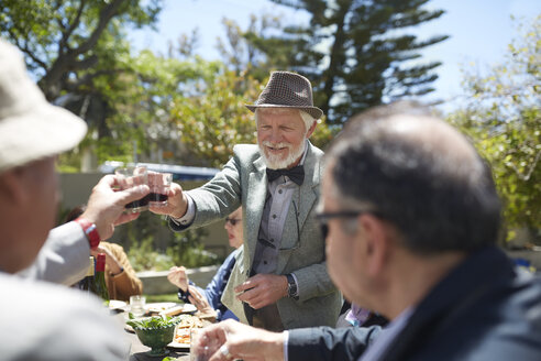 Älterer Mann in Anzug und Fliege stößt mit Freunden auf einer sonnigen Gartenparty mit Wein an - CAIF20734