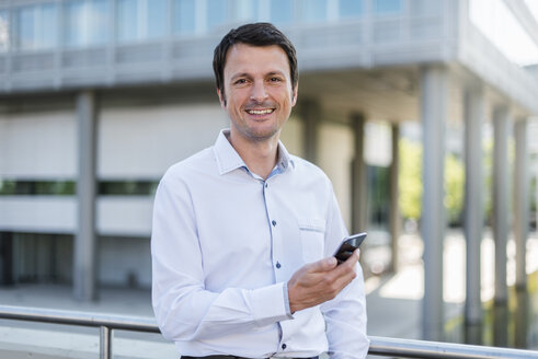 Porträt eines lächelnden Geschäftsmannes mit Mobiltelefon in der Stadt - DIGF04654