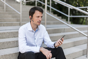 Businessman with headset and cellphone sitting on stairs - DIGF04678