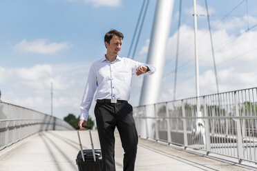 Businessman with rolling suitcase on a bridge checking the time - DIGF04681